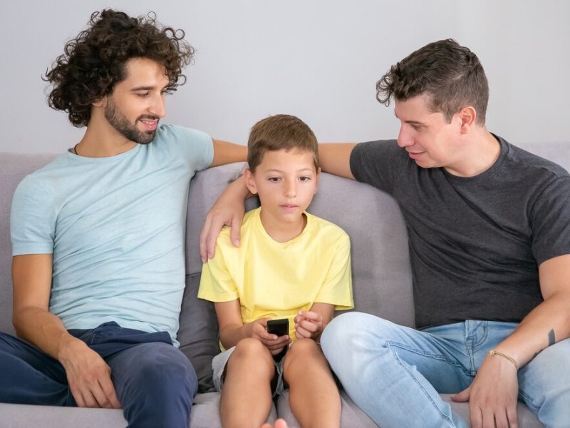pais-gays-positivos-e-filho-assistindo-programa-de-tv-juntos-em-casa-sentados-no-sofa-da-sala-sorrindo-e-abracando-o-menino-conceito-de-entretenimento-familiar-e-domestico_74855-11919